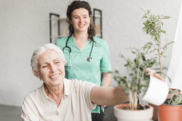 nurse-looking-senior-woman-sitting-wheelchair-watering-plant